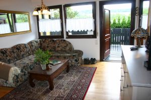 Auch durch die große Fensterfront des Wohnzimmers genießen Sie die Fernsicht. Eine komfortable Sitzecke (m. Schlaffunktion) lädt zum gemütlichen Erzählen, Planung von Ausflügen oder einfach zum Entspannen ein. - Bild 7: Idyll. Ferienhaus EifelNest & uneinsehb. Sonnenterrasse mit Fernblick